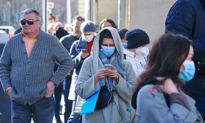 Женщинна идёт в толпе людей в медицинской маске и смотрит в телефон